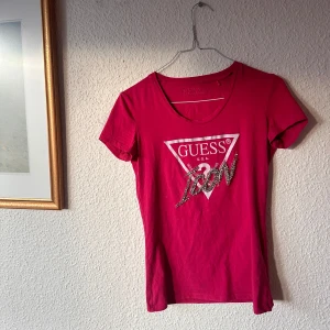 Röd t-shirt från Guess - Snygg röd t-shirt från Guess (vet inte om den är äkta)  med det ikoniska logotyptrycket i silver och vitt på framsidan. Den har en v-ringad hals och korta ärmar, perfekt för en casual look. 