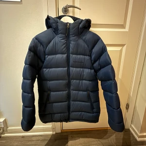 Mörkblå dunjacka från Peak Performance - Säljer en snygg mörkblå dunjacka från Peak Performance. Jackan har en dragkedja framtill och en praktisk huva. Perfekt för kyliga dagar med sin vadderade design.