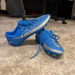 Blå Nike fotbollsskor - Snygga blå Nike fotbollsskor med vita detaljer och snörning. Skorna har gröna dobbar för bra grepp på planen. Perfekta för fotbollsträning och matcher. Använt skick men bra skick.