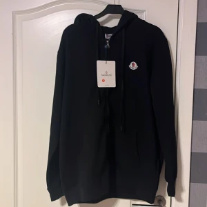 Svart hoodie från Moncler - Säljer en snygg svart hoodie från Moncler i nytt skick. Den har en klassisk design med Moncler-loggan på bröstet och en justerbar huva. Perfekt för både höst och vinter, och passar till alla tillfällen. Skön och stilren, ett måste i garderoben! 🖤