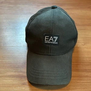 Svart keps från Emporio Armani EA7 - Snygg svart keps från Emporio Armani EA7. Den har en broderad logga framtill och justerbart spänne bak för perfekt passform. Kepsen är i ganska dåligt skick men funkar 