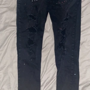 Svarta jeans från Dsquared2 - Säljer ett par svarta jeans från Dsquared2 i bra skick. De har en cool distressed look med slitningar och färgstänk. Tillverkade i Italien av 98% bomull och 2% elastan för en bekväm passform. Perfekta för en avslappnad och trendig stil!