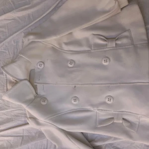 Vit kappa med rosetter - Elegant vit kappa med knappar och dekorativa rosetter framtill. Jackan har trekvarts ärmar och en klassisk krage, perfekt för en stilren look.