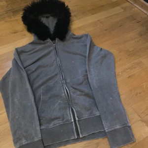 Grå hoodie med pälshuva - Säljer en grå hoodie med dragkedja och en mysig pälskantad huva. Jackan har en avslappnad passform. Den är i bra skick men har lite slitage som ger den en cool vintage-look. Passar herr L