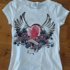 Vit t-shirt med tryck - Säljer en cool vit t-shirt med ett stort tryck av vingar, rosor och ett hjärta i mitten. Perfekt för en avslappnad stil eller en dag ute med vänner. T-shirten är i bra skick och har korta ärmar. Passar perfekt till jeans eller shorts!