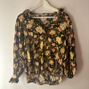 Blus Carin Wester - Snygg blus med blommigt mönster i grönt, rosa och gult. Blusen har en krage med volang och en knäppning framtill. Ärmarna är  långa och har en volang vid handleden. Blusen är tillverkad i ett tunt och lätt material. Storlek 40 / L