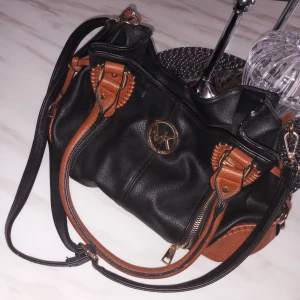 Mikael kors Svart och brun axelväska med dragkedja - Snygg svart axelväska i skinn med bruna detaljer och dragkedjor. Perfekt för vardagsbruk med gott om plats för dina saker. Väskan har ett elegant märke framtill och justerbar axelrem för bekväm passform.