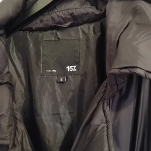 Svart lång kappa från 157 - Säljer en svart lång kappa från 157 i storlek S. Den är perfekt för höst och vinter med sitt långa snitt och varma material. Kappan har långa ärmar och knappar framtill. Den är i mycket bra skick och redo för nya äventyr!