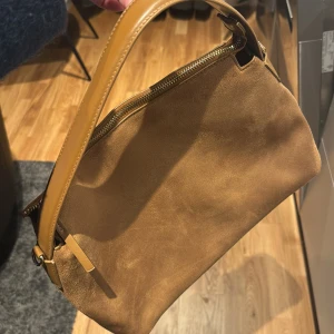 Brun axelväska från H&M - Snygg brun axelväska från H&M i mockaimitation. Jsterbar axelrem som enkelt kan ta av och dragkedja upptill. Liten defekt kom så när jag fick hem den. Helt oanvänd 