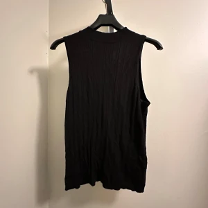Svart ärmlös topp - Snygg svart ärmlös topp i ribbat material. Perfekt för både vardag och fest. Den har en hög hals och är i ett mjukt och stretchigt tyg som ger en bekväm passform. Helt oanvänd från Kappahl i storlek M!