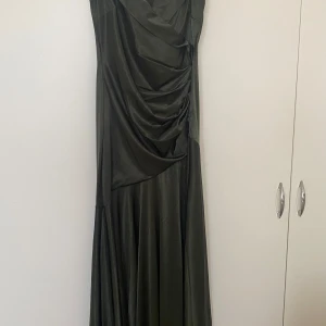 Grön långklänning med slits - Snygg grön långklänning med en elegant slits framtill. Klänningen har en draperad design och offshoulder-stil som ger en lyxig känsla. Perfekt för festliga tillfällen! Endast använd 1 gång
