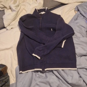 Mörkblå kofta från Ysl - Säljer en snygg mörkblå kofta från ysl i bra skick. Den har en dragkedja framtill och vita ribbade detaljer vid ärmslut och nederkant. Perfekt för höst och vår, och funkar både till vardags och lite finare tillfällen. Stor logga på bröstet ger en klassisk touch.