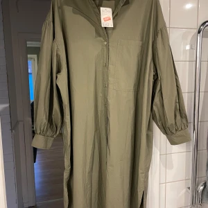 Olivgrön långklänning med knappar - Säljer en olivgrön långklänning i nytt skick. Den har långa ärmar med knappar vid ärmsluten och en bröstficka. Perfekt för höst och vår med sin avslappnade passform och stilrena design.