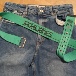 Grönt unikt bälte från Jack & Jones - Snyggt grönt bälte från Jack & Jones med texten 'JEANS INTELLIGENCE'. Bältet har en silverfärgad spänne och flera justerbara hål för perfekt passform. Perfekt för att ge en färgklick till din outfit!