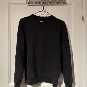 Svart tröja från C.P. Company - Snygg svart tröja från C.P. Company med rund hals och långa ärmar. Tröjan har en unik detalj på ärmen med en liten ficka och C.P. Company-logga. Liten skada på Cp Company patchen. 