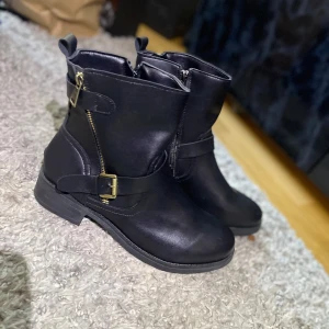 Svarta boots med dragkedja och spänne - Snygga svarta boots i skinn med dragkedja och dekorativt spänne. Perfekta för höst och vinter med en stilren design och låg klack. Helt oanvända pga fel storlek 