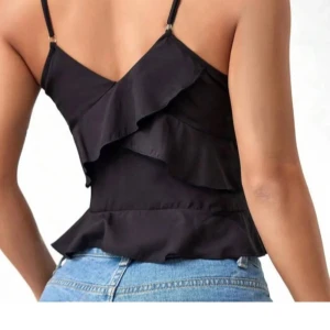 Svart volangtopp - Snygg svart topp med volangdetaljer och smala axelband. Perfekt för fest, helt oanvänd köpte i fel färg