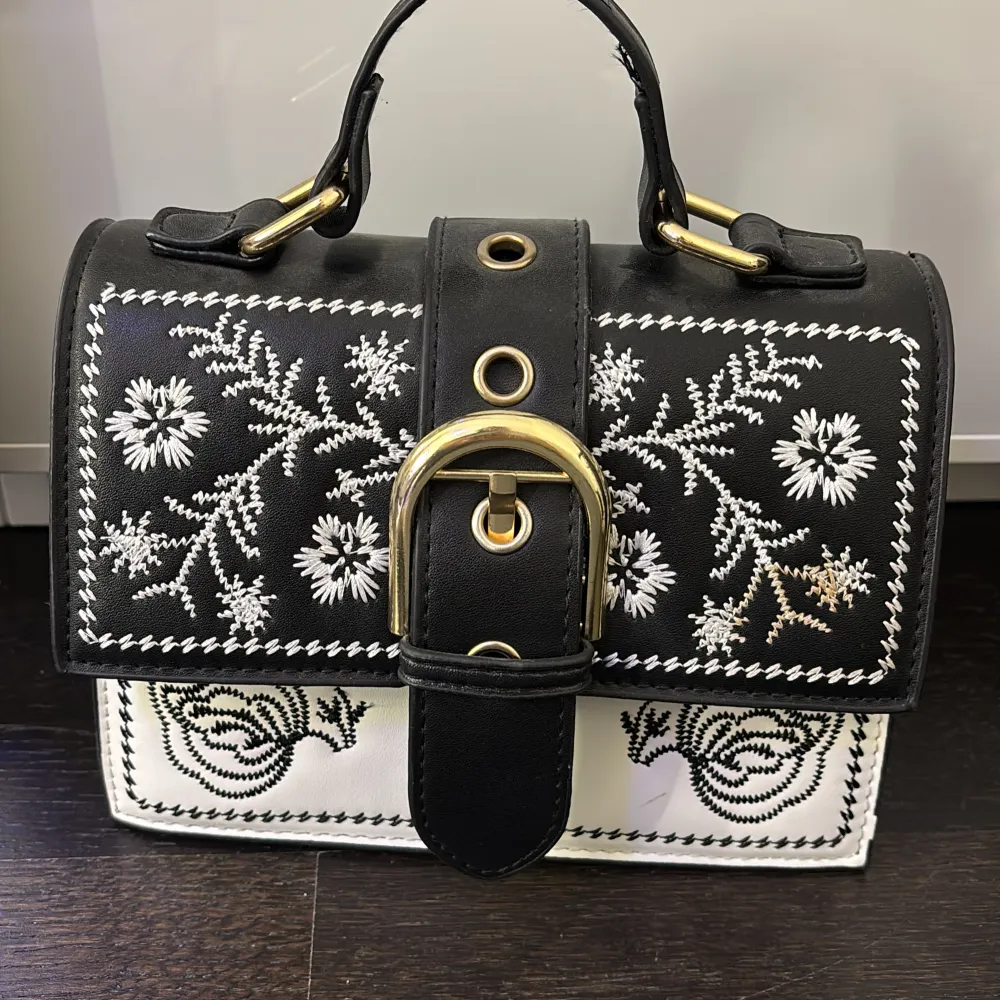 Snygg handväska i svart och vitt med vackra broderade blommor. Väskan har en stor guldfärgad spänne och ett praktiskt handtag. Perfekt för att ge en elegant touch till din outfit!. Laukut & Käsilaukut.