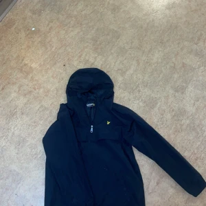 Svart vindjacka från Lyle & Scott - Säljer en svart vindjacka från Lyle & Scott i bra skick. Jackan har en dragkedja framtill och en liten gul logga på bröstet. Perfekt för höst och vår, med en stilren look som funkar till det mesta. Den har långa ärmar och en huva för extra skydd mot väder och vind. priset kan diskuteras