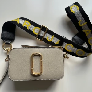 Beige axelväska från Marc Jacobs - Snygg beige axelväska från Marc Jacobs i mycket bra skick. Väskan har en justerbar rem, logga i gult och silver. Praktisk dragkedja och en stilren design med gulddetaljer. Perfekt för både vardag och fest! Mindre slitage (se sista bilden) dock inget som man tänker på. 