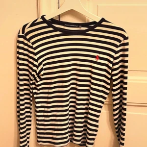 Randig långärmad tröja från Ralph Lauren - Säljer en snygg och klassisk randig tröja från Ralph Lauren i storlek M. Tröjan är mörkblå och vit med långa ärmar och har det ikoniska röda Ralph Lauren-märket på bröstet. Perfekt för både vardag och lite mer uppklädda tillfällen. 🖤🤍
