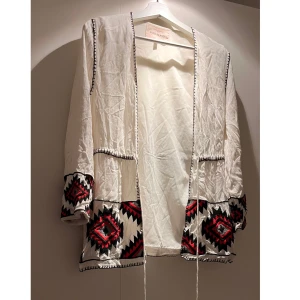 Vit kimono från River Island - Säljer en superfin vit kimono från River Island med snyggt broderat mönster i svart och rött. Den har långa ärmar och knytband framtill. Säljs pga för liten tyvärr 