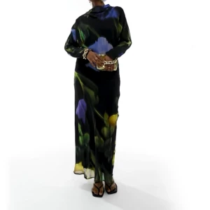 Blommig långklänning i mörka toner - Säljer en elegant långklänning med ett vackert blommönster i blått, gult och grönt på en svart bakgrund. Klänningen har långa ärmar och en lös passform, perfekt för höst- och vårsäsongen. Materialet är lätt och flödande, vilket ger en sofistikerad look. Perfekt för festliga tillfällen!