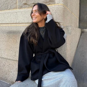 Missäy kappa - Säljer en missäy elegant svart kappa med knytband i midjan. Perfekt för höst och vår, med långa ärmar och en stilren design. Passar både till vardags och festliga tillfällen. Den är i bra skick.