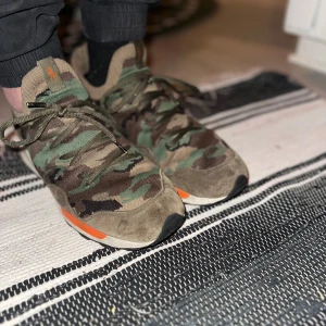 Camouflage sneakers från ralph lauren - Säljer ett par coola sneakers från ralph lauren  i camouflage-mönster. De har en snygg kombination av gröna och bruna nyanser med orange detaljer. Skorna har snörning och är perfekta för en avslappnad stil. De är i bra skick och redo för nya äventyr!