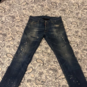 Blå jeans från Dsquared2 - Säljer ett par coola blå jeans från Dsquared2 i storlek 48. De har en unik distressed look med slitningar och färgstänk som ger en edgy stil. Perfekta för en avslappnad men trendig outfit. Tillverkade i Italien, vilket garanterar hög kvalitet.