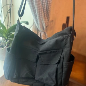 Svart axelväska med fickor - Säljer en praktisk svart axelväska i syntetmaterial. Väskan har två fickor framtill och en dragkedja upptill för säker förvaring. Perfekt för vardagsbruk och rymmer allt du behöver ha med dig. Justerbar axelrem för bekväm passform.