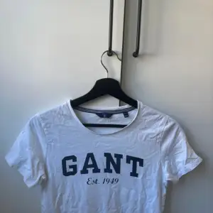 Säljer en vit t-shirt från GANT i storlek XS. Den har en klassisk design med GANT-loggan och 'Est. 1949' tryckt i blått på framsidan. Perfekt för en avslappnad stil. T-shirten är i bomull och har korta ärmar.