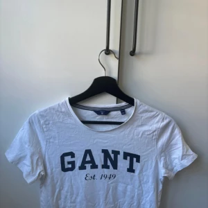 Vit t-shirt från GANT - Säljer en vit t-shirt från GANT i storlek XS. Den har en klassisk design med GANT-loggan och 'Est. 1949' tryckt i blått på framsidan. Perfekt för en avslappnad stil. T-shirten är i bomull och har korta ärmar.
