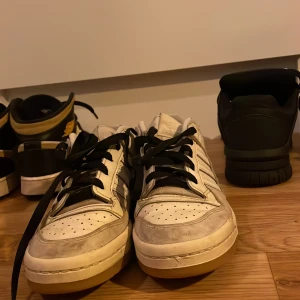 Vita Adidas sneakers med svarta detaljer - Säljer ett par vita Adidas sneakers i storlek 38. De har svarta snören och klassiska ränder på sidan. Skorna är lite smutsiga men i bra skick. Perfekta för vardagsbruk eller en avslappnad stil. Passar bra till jeans eller shorts!