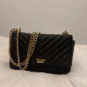 Svart quiltad axelväska från Victoria's Secret - Snygg svart axelväska från Victoria's Secret i quiltat mönster. Väskan har en elegant kedjerem i guld och ett stilrent innerfoder med ränder. Perfekt för både vardag och fest! Den är lite mer använd därav priset