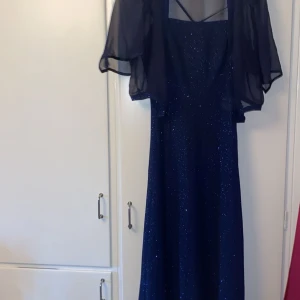 Mörkblå glittrig långklänning - Säljer en elegant mörkblå långklänning med glittrande detaljer. Klänningen har ett transparent cape-liknande överdrag och en stilren design. Perfekt för festliga tillfällen eller nyårsfirande. Den är i nyskick och redo att bäras av någon som vill glänsa!