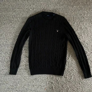 En svart flätad gant tröja med märke i fram på bröstet  - Det är en small tröja som är väldigt bra skick och är väldigt stilig. Pris kan diskuteras