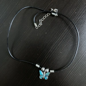 Kort halsband med turkos fjäril, berlock, silver, hänge, svart, y2k, 90 tal, 90s - Halsband med fjäril, det svarta plastsnöret är ganska stelt. Finns missfärgningar på metallen. Längd: 45-51 cm, hänge bredd: 1,9 cm.