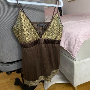 Brun paljettprydd topp från Vero Moda  - Snygg brun topp från Zara med tunna axelband och glittrande paljetter i guld på bysten och nederkanten. Perfekt för en kväll ute eller en festlig tillställning. Toppen har en v-ringad design och är tillverkad i ett lätt och bekvämt material.