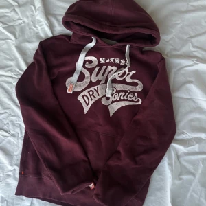 Vinröd hoodie från Superdry - Säljer en snygg vinröd hoodie från Superdry med tryck framtill. Den har en justerbar huva med vita snören och ribbade muddar vid ärmslut och nederkant. Perfekt för en avslappnad stil.