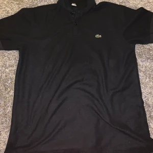 Svart kortärmad pikétröja från Lacoste - Säljer en klassisk svart pikétröja från Lacoste med korta ärmar och den ikoniska krokodilloggan på bröstet. Perfekt för en stilren och avslappnad look. Tröjan har en knappslå framtill.