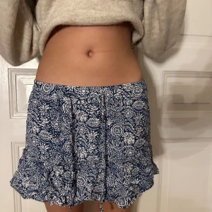 Blå blommig kjol med volang - Säljer en söt blå kortkjol med vitt blommönster och volangdetaljer. Kjolen har en bekväm passform och knytband i midjan för justering. Perfekt för en somrig look! Från Zara. Skrynklig men går lätt att fixa, köpare står för frakt 