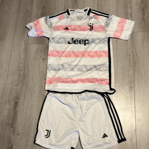 Juventus fotbollströja och shorts - Säljer en Juventus fotbollströja och shorts från Adidas. Tröjan är kortärmad med ett unikt mönster i vitt, rosa och grått, samt svarta detaljer. Shortsen är vita med svarta ränder på sidorna. Perfekt för fotbollsfans som vill ha en stilren look på planen.Byxorna har även en lite fläck på sig.