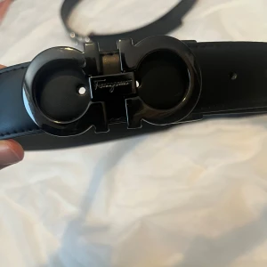 Ferregamo bälte svart - Snyggt svart bälte från Ferragamo med ett stilrent metallspänne i svart. Perfekt för att ge en elegant touch till din outfit. Bältet är tillverkat i skinn och har en klassisk design som passar till många olika stilar.