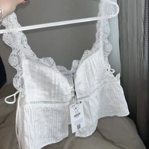 Vit spetsprydd topp från Bershka - Säljer en söt vit topp från Bershka med spetsdetaljer och knytband i sidorna. Sprillans ny och oanvänd!