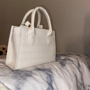 Vit handväska med krokodilmönster - Snygg vit handväska med krokodilmönster och två handtag. Perfekt för att ge en stilren touch till din outfit. Väskan har en strukturerad design som ger den ett elegant utseende, finns även axelrems band så man kan hänga den över sig!🤗aldrig använd
