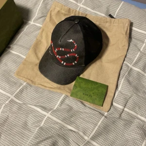 Svart keps med ormmönster - Snygg svart keps med ett iögonfallande ormmönster i rött och vitt framtill. Perfekt för att ge din outfit en cool touch. Kepsen har en klassisk passform och justerbar rem baktill.