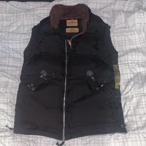 Svart dunväst från Parajumpers - Säljer en svart dunväst från Parajumpers med brun krage och dragkedja framtill. Västen har flera praktiska fickor och en broderad logga på baksidan. Perfekt för kyliga dagar.