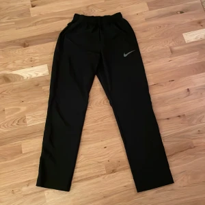 Svarta träningsbyxor från Nike - Säljer ett par svarta träningsbyxor från Nike med Dri-FIT-teknologi. Byxorna har en elastisk midja och en diskret Nike-logga på benet. Perfekta för träning och bekväma att bära. 