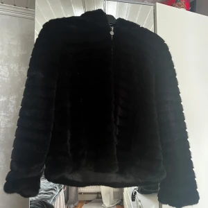 Svart pälsjacka från Flash Woman - Snygg svart pälsjacka från Flash Woman i storlek M. Jackan har en dragkedja framtill samt en luva  och är perfekt för kyligare dagar. Den är mjuk och har en lyxig känsla med sitt pälsmaterial.